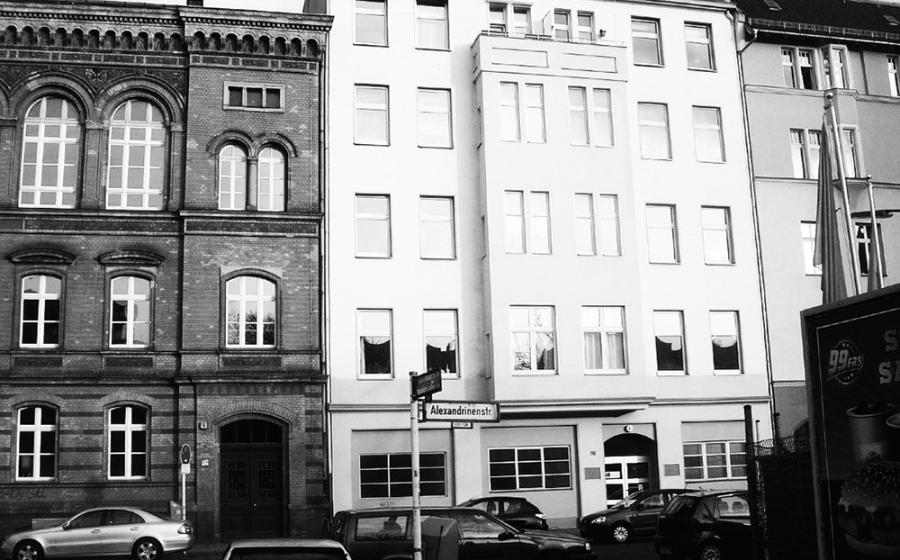 Black and white photo with a street sign for Alexandrinenstraße in the foreground, with one white residential buidling in its entirety behind the sign, and a sliver of another residential building to the right and a larger chunk of a dark brick (former factory? school?) building to the left. 
