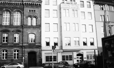 Black and white photo with a street sign for Alexandrinenstraße in the foreground, with one white residential buidling in its entirety behind the sign, and a sliver of another residential building to the right and a larger chunk of a dark brick (former factory? school?) building to the left. 